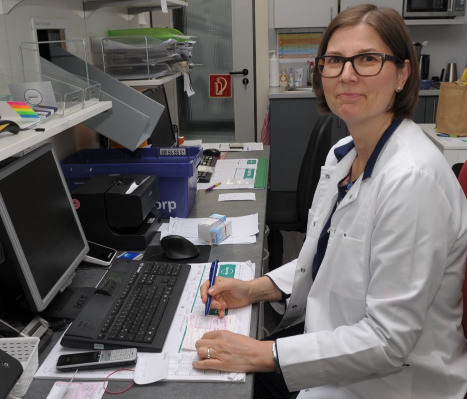 Eine Mitarbeiterin der Babelsberg Apotheke in Potsdam sitzt am Schreibtisch, bearbeitet Rezepte und schaut lächelnd in die Kamera.